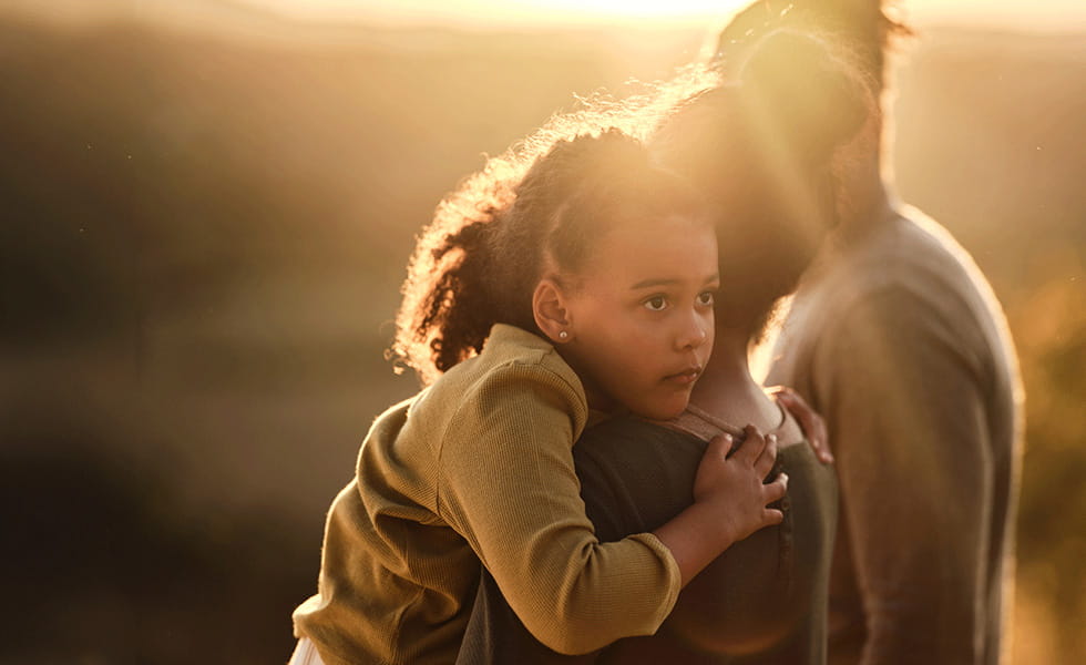 A mother and young daughter sit at the top of their garden steps, facing away from camera. The mother appears to be comforting the daughter.