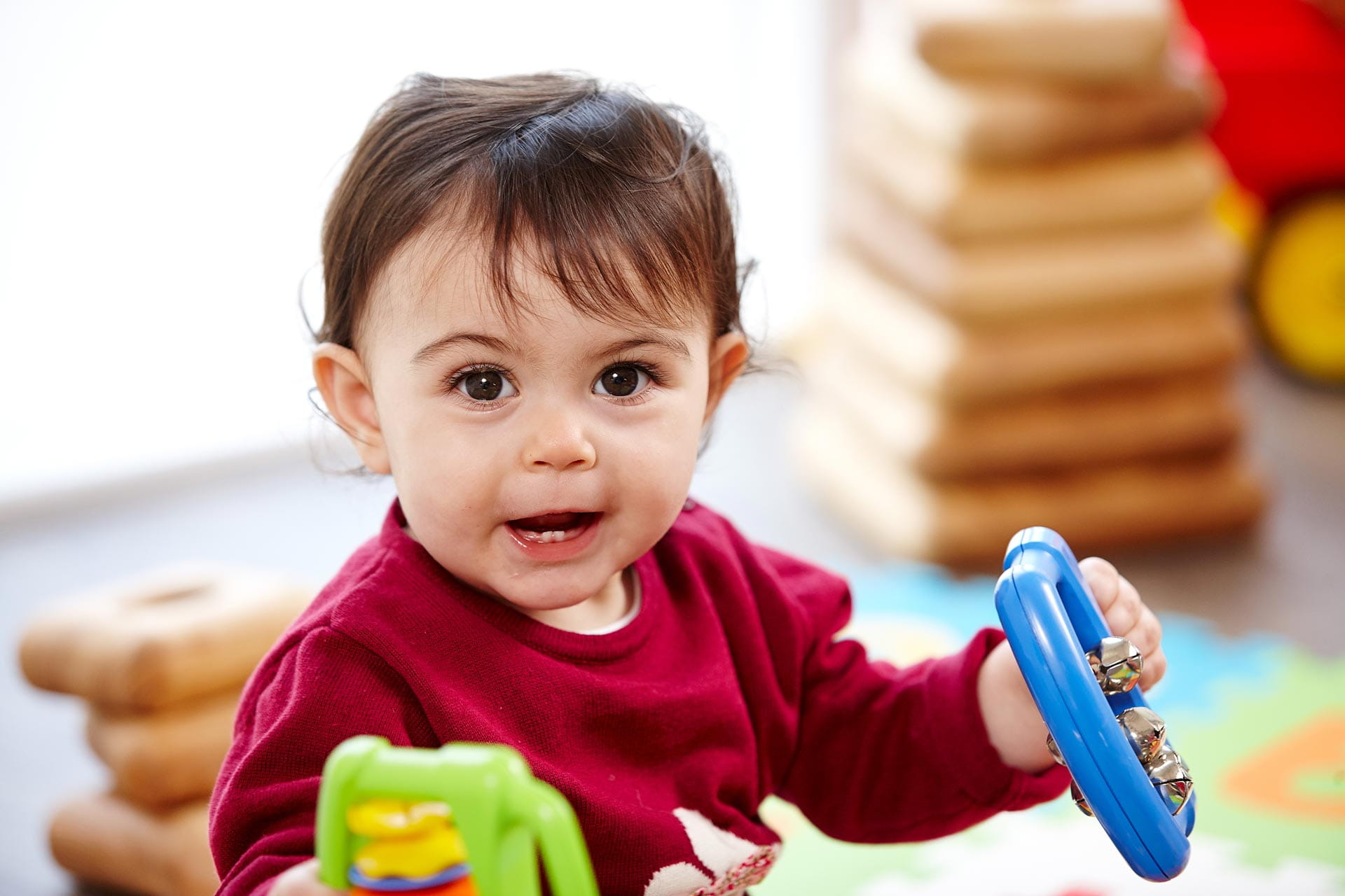 A baby is smiling whilst playing with a rattle in each hand