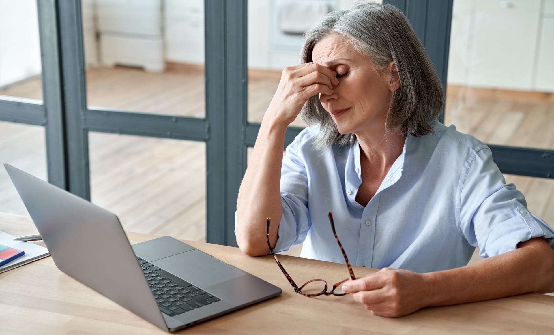 Tired older lady sitting at her desk holding glasses and rubbing her eyes